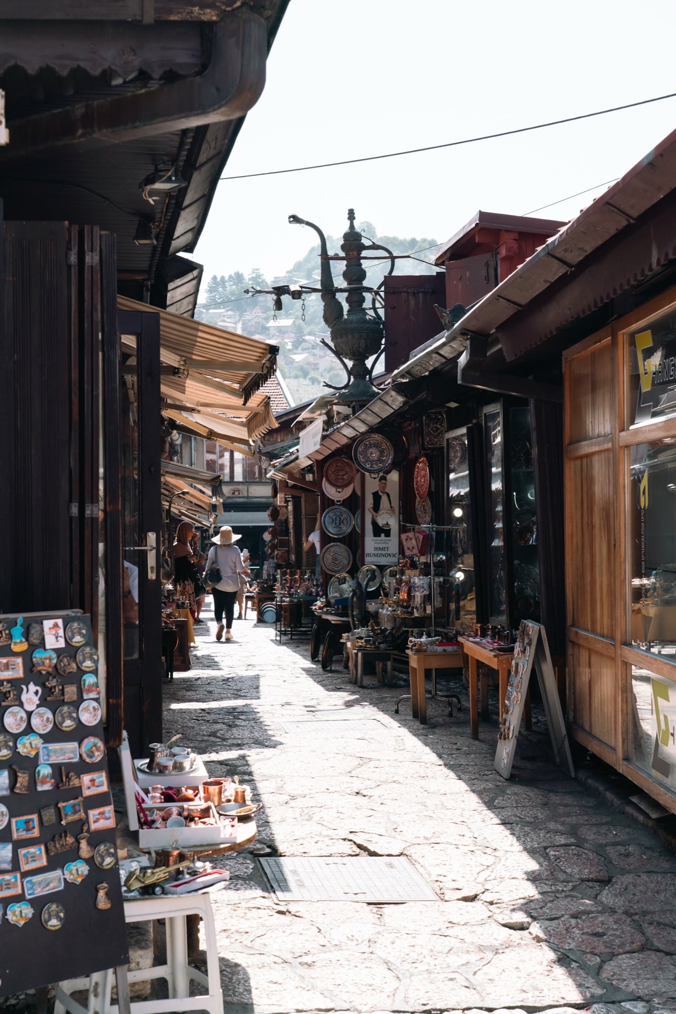 The Kazandžiluk coppersmiths' alley in Baščaršija, a giant metal jug sign overhead and tables of hammered copperware and souvenir magnets lining the shopfronts