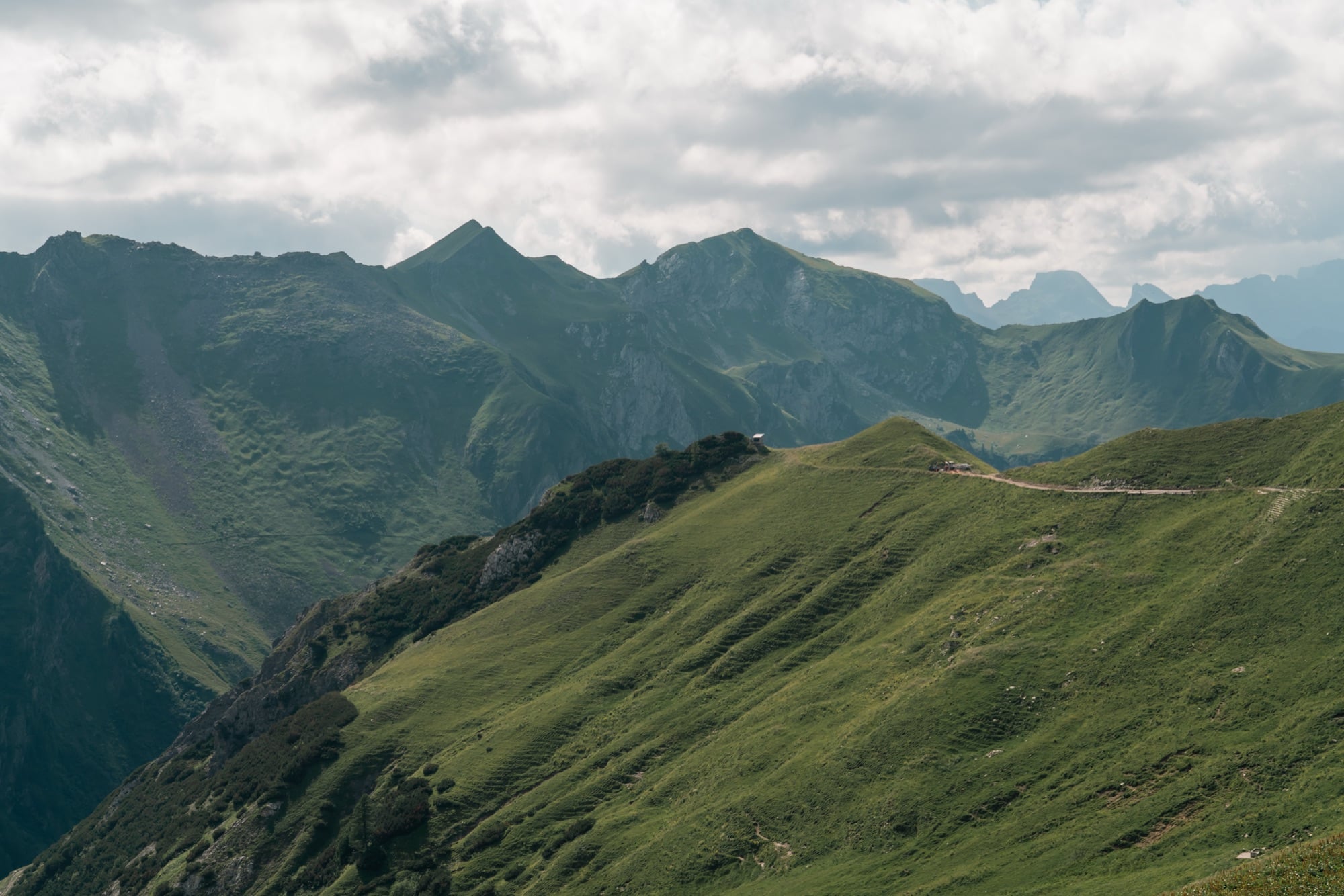 Rolling green alpine ridges with a footpath tracing a grassy crest, jagged peaks hazy in the distance