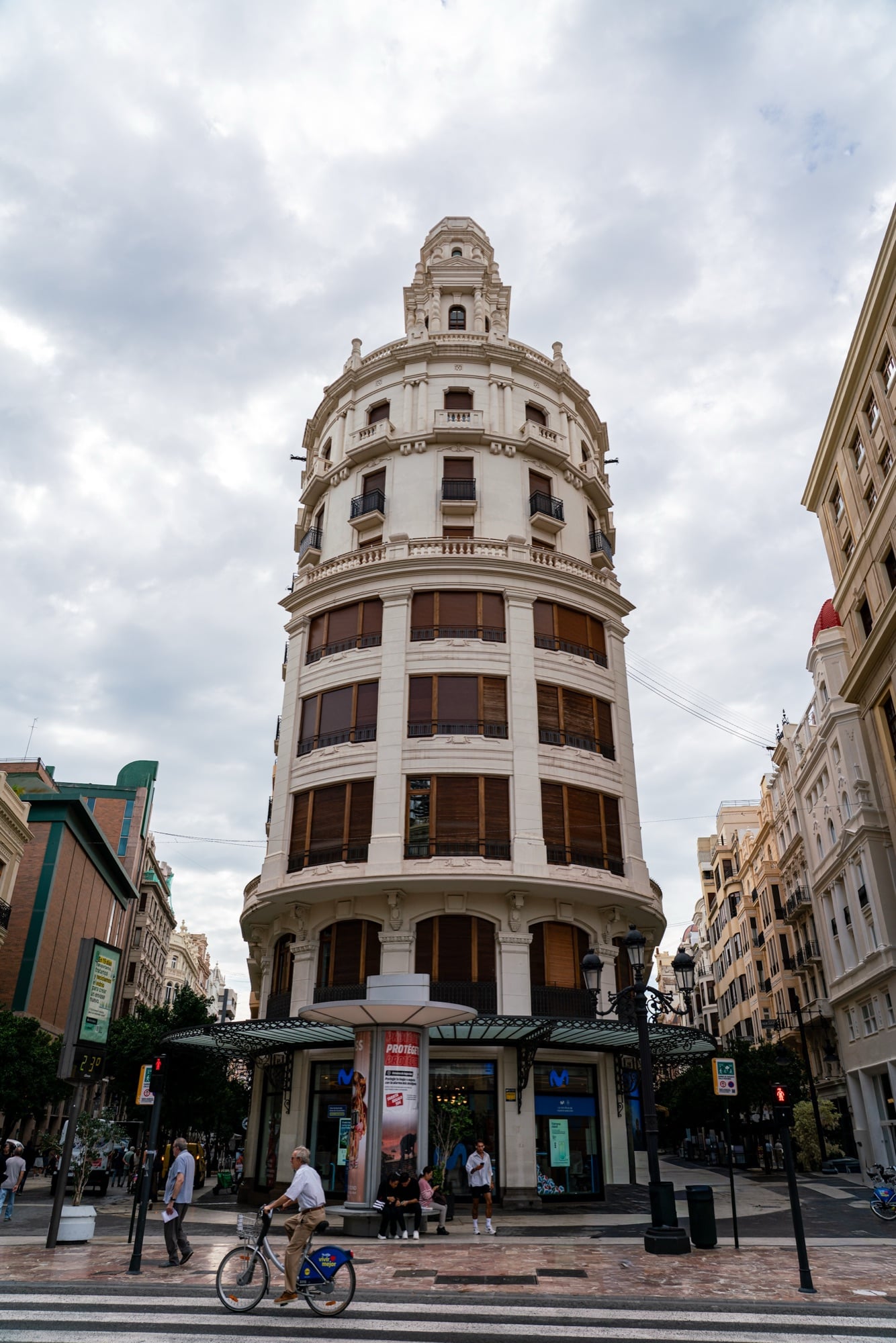 A round-cornered tower building with banded windows rising over a busy intersection, a cyclist crossing in front