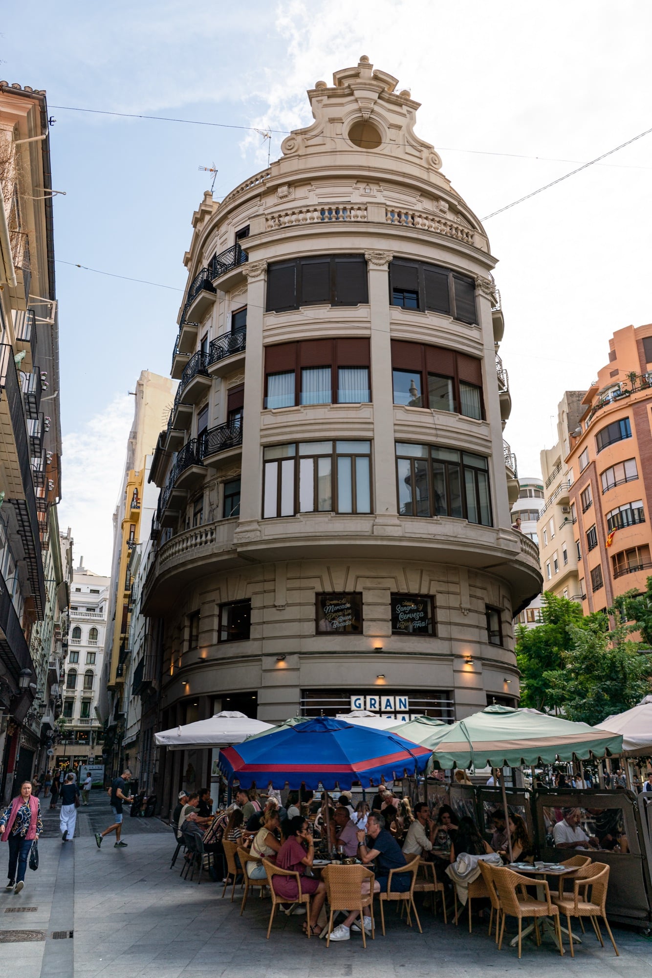 A curved belle-époque corner building above a crowded café terrace of striped umbrellas