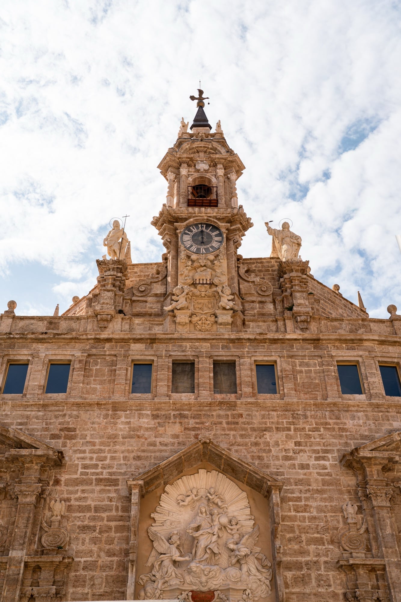 A sandstone Baroque church facade topped by a clock and sculpted figures against a white sky