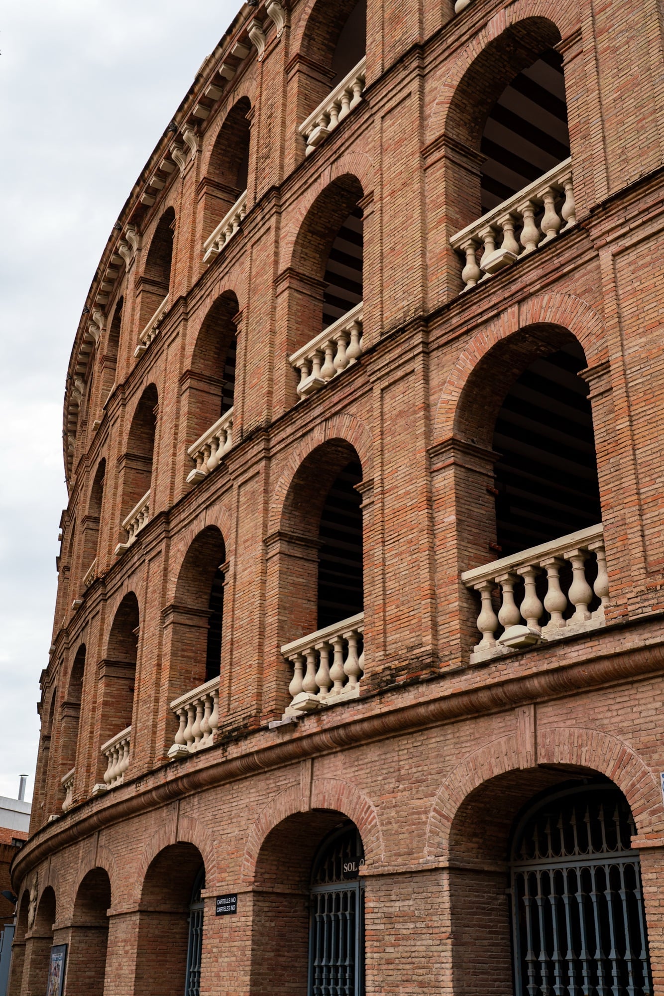 The curving red-brick wall of Valencia's Plaza de Toros, tiers of arches lined with cream stone balustrades rising toward a pale sky